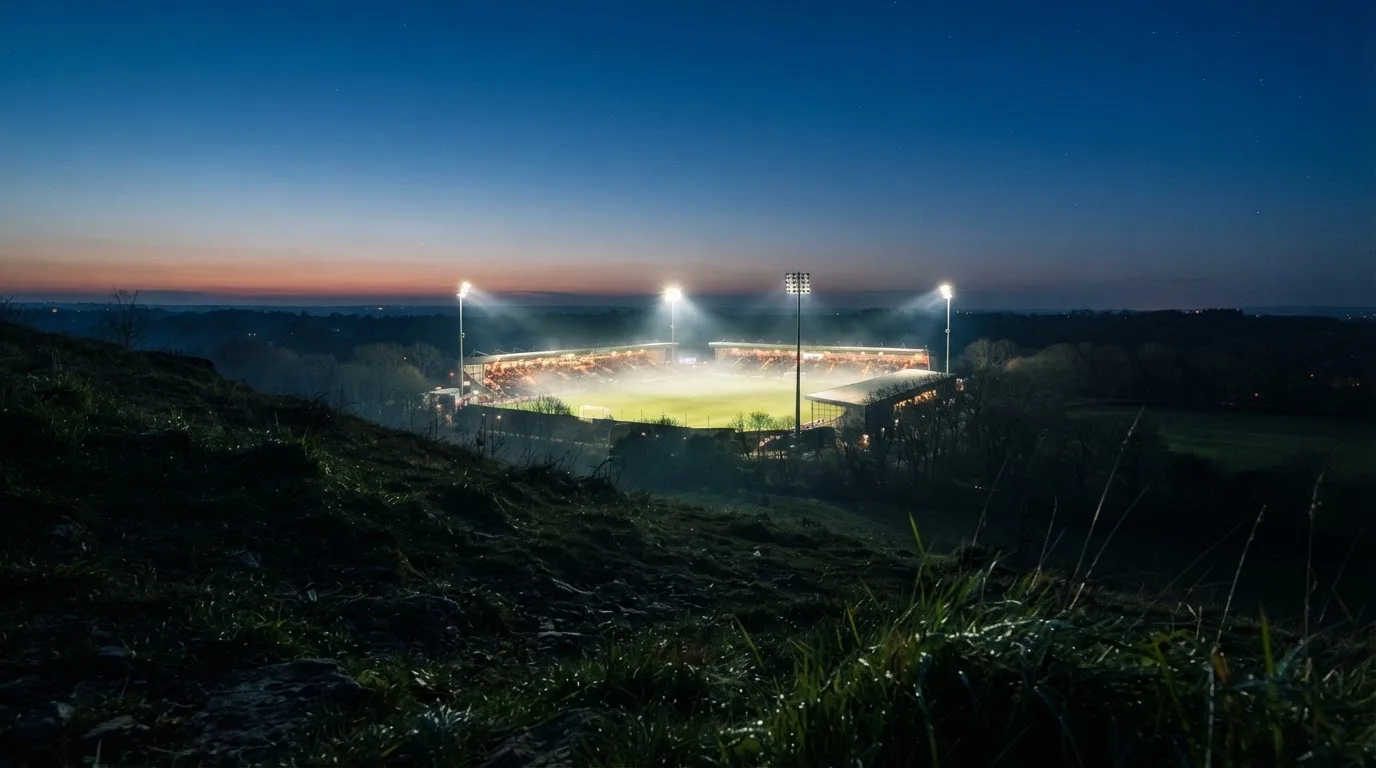 Ein Stadion unter Flutlicht bei Nacht, von einem Hügel aus der Ferne betrachtet, mit Dunst über dem Spielfeld.