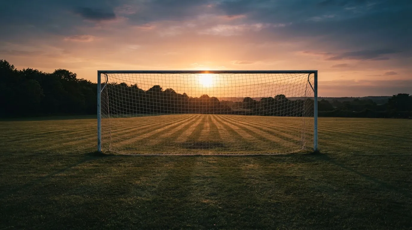Ein Fußballtor mit Netz bei Sonnenuntergang auf einem leeren Platz, die Schatten der Torpfosten liegen lang auf dem Gras.