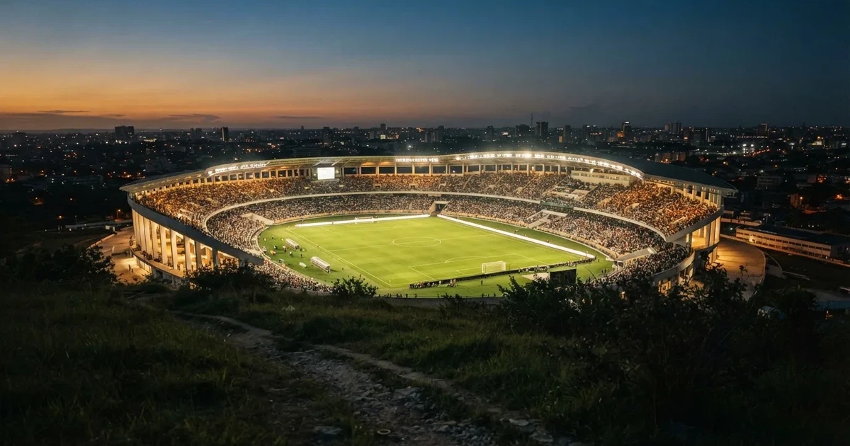 Ein Fußballstadion unter Flutlicht mit hellen Rängen, von einem Hügel aus gesehen, darüber ein dunkelblauer Abendhimmel.