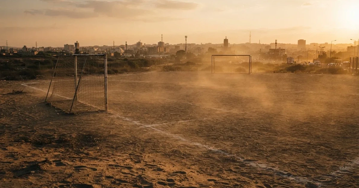 Ein sandiger Fußballplatz am Rand einer Stadt in warmem Abendlicht, Staub in der Luft und zwei Tore in der Ferne.