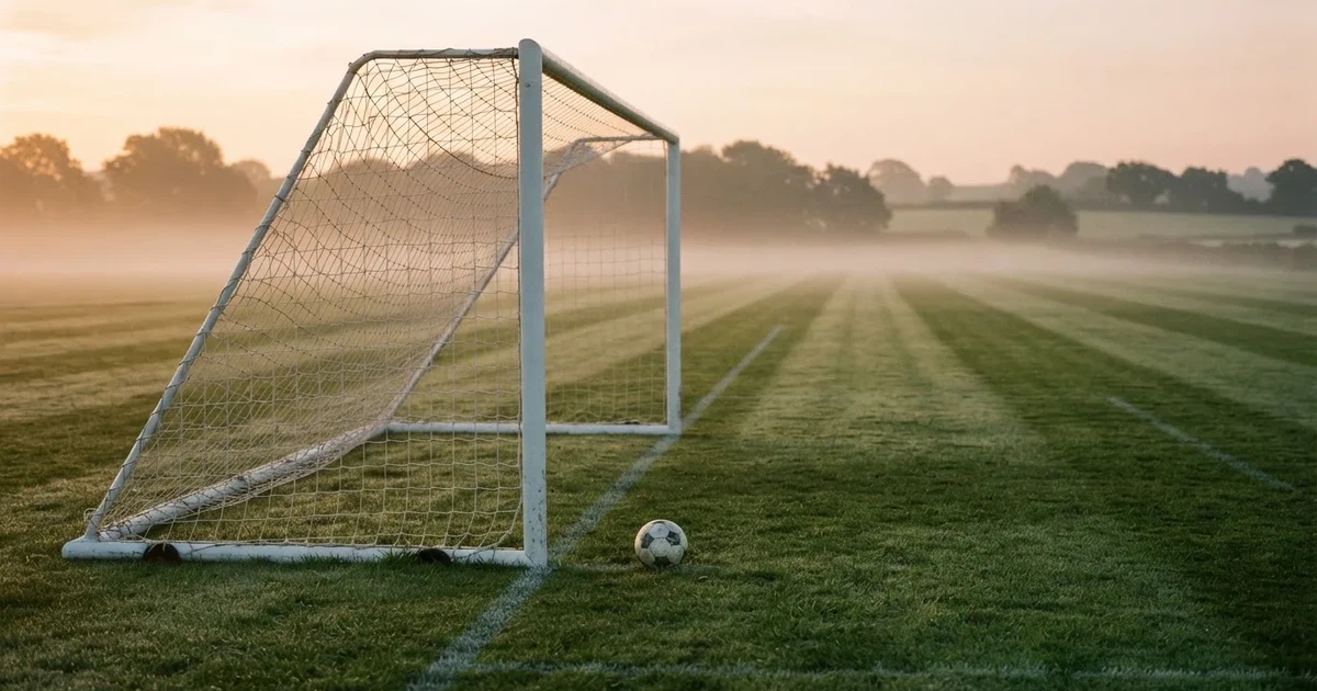 Ein Fußballtor mit gespanntem Netz steht auf einem leeren Rasenplatz, davor ein einzelner Ball, Morgendunst liegt über dem Feld.
