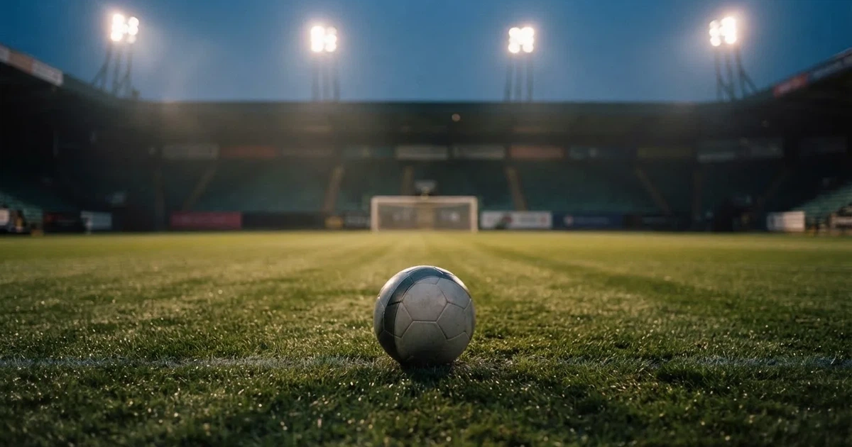 Ein Fußball liegt auf dem Rasen eines leeren Stadions bei Flutlicht, im Hintergrund unscharfe Tribünen ohne erkennbare Beschriftung.