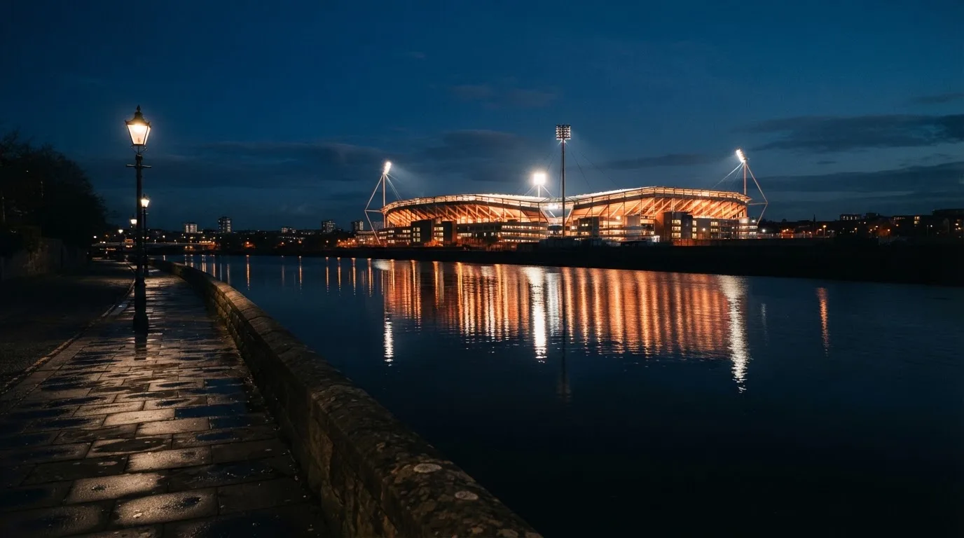 Ein Stadion unter Flutlicht aus der Ferne gesehen, im Vordergrund ein ruhiger Fluss und ein Fußweg.