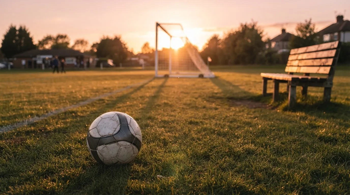 Ein Fußball auf einem Rasenplatz im Gegenlicht der tiefstehenden Sonne, am Rand eine leere Ersatzbank.