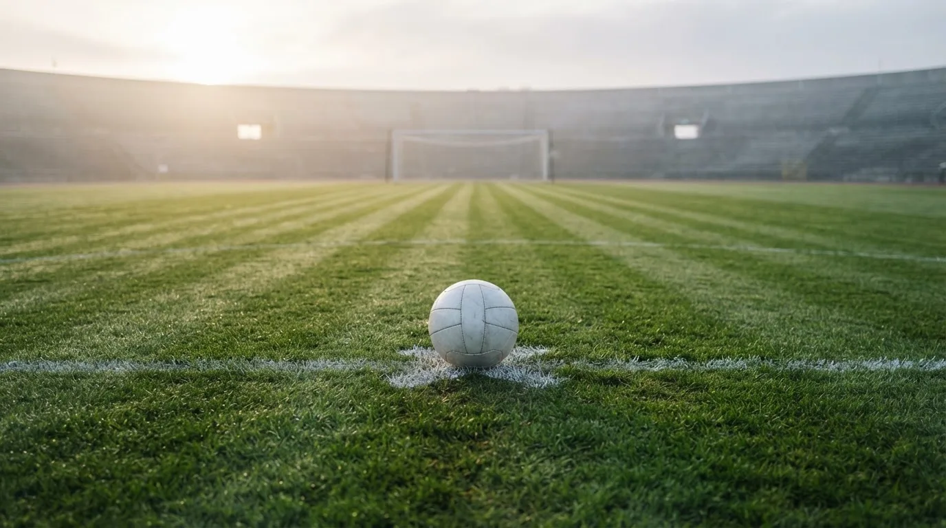 Ein Fußball liegt auf dem Anstoßpunkt eines leeren Rasenplatzes, im Hintergrund hängt Morgendunst über den Tribünen.