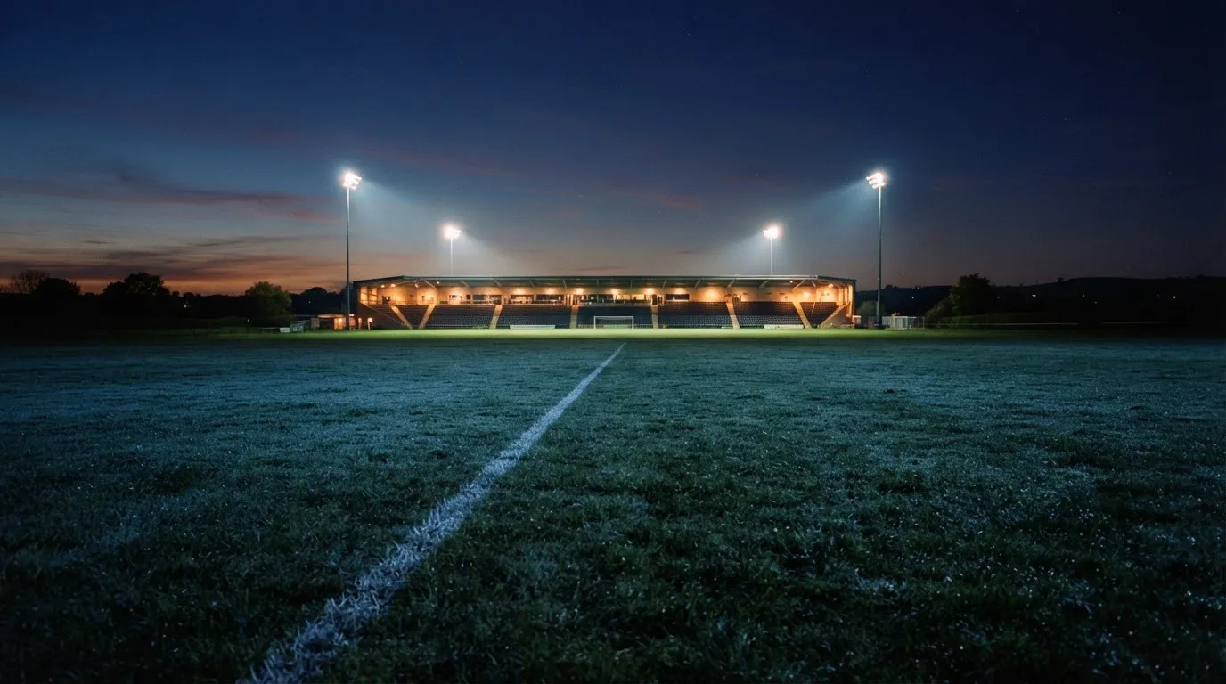 Ein Stadion unter Flutlicht aus der Ferne, davor ein stilles Feld im Dämmerlicht.