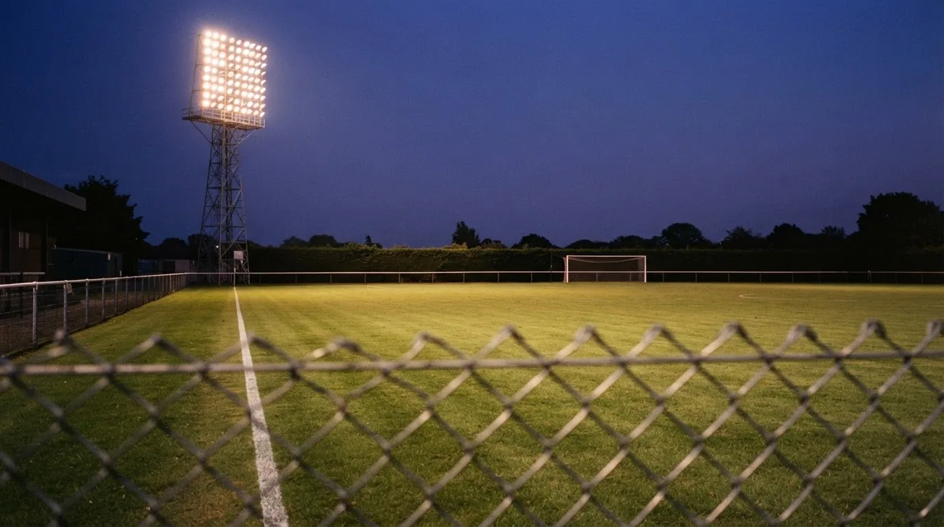 Eine Flutlichtanlage über einem leeren Sportplatz am Abend, im Vordergrund ein Maschendrahtzaun.