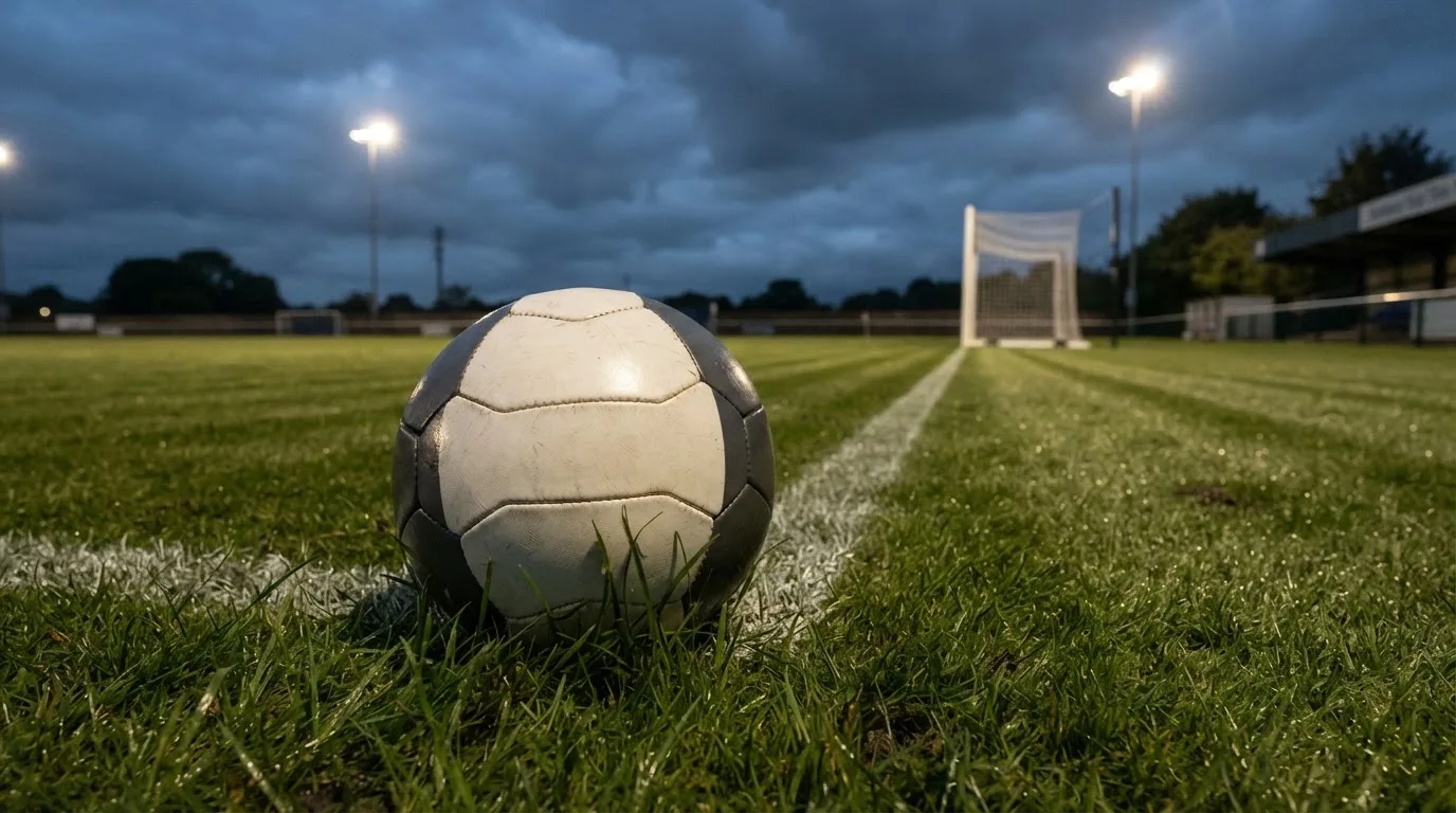 Ein Fußball auf einem Rasenplatz, im Hintergrund Flutlichtmasten und ein bewölkter Himmel.