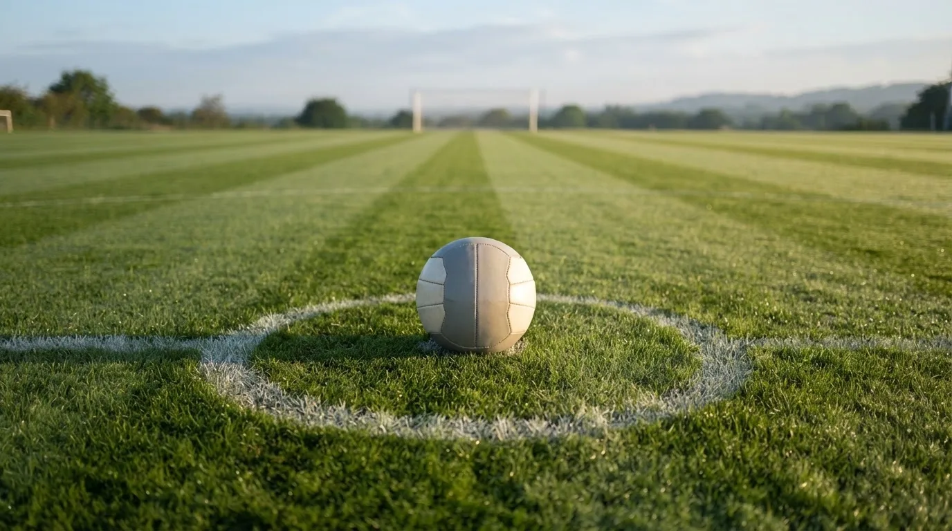 Ein Fußball auf dem Mittelkreis eines leeren Rasenplatzes im frühen Morgenlicht.