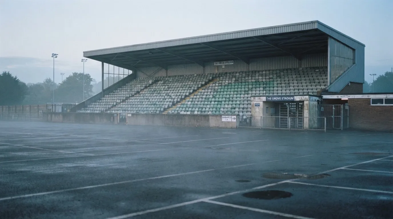 Eine leere Tribüne eines kleinen Stadions am frühen Morgen, aus der Ferne über einen menschenleeren Parkplatz fotografiert.