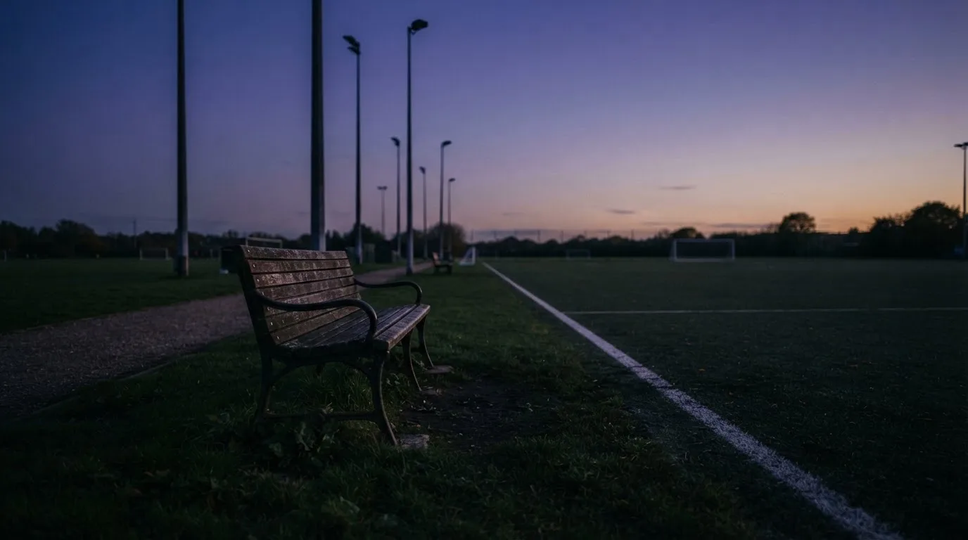 Eine leere Parkbank am Rand eines Fußballfelds in der Abenddämmerung, im Hintergrund ragen dunkle Flutlichtmasten in den Himmel.