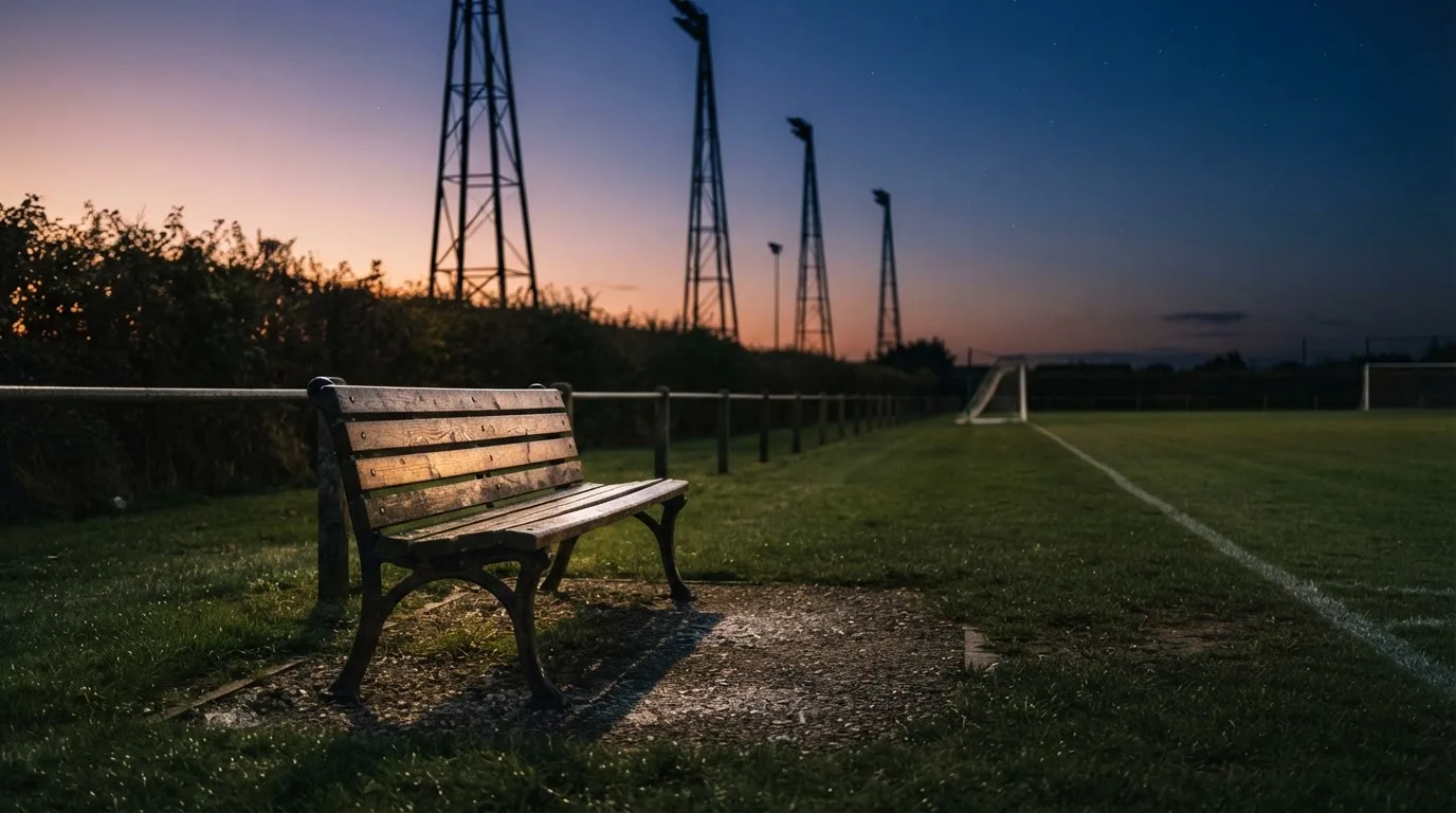 Eine leere Parkbank am Rand eines Fußballfelds in der Abenddämmerung, im Hintergrund ragen dunkle Flutlichtmasten in den Himmel.