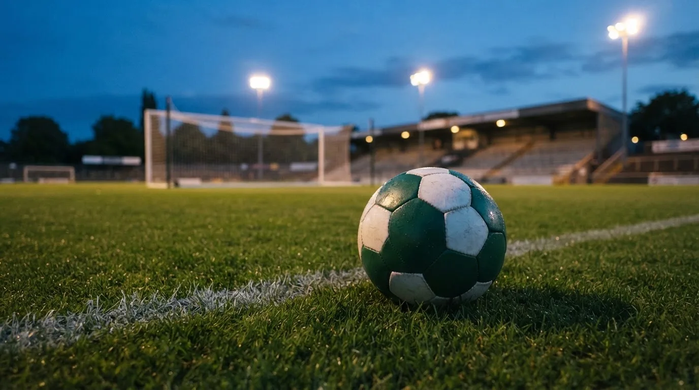 Ein Fußball liegt auf dem Rasen eines leeren Platzes, im Hintergrund ein Stadion unter Flutlicht in der Abenddämmerung.