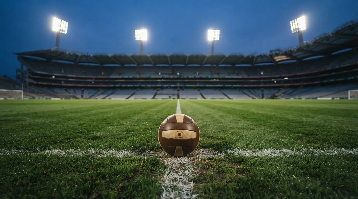 Ein Fußball liegt auf dem Anstoßpunkt eines leeren Rasenplatzes, im Hintergrund steht ein Stadion unter Flutlicht in der Dämmerung.