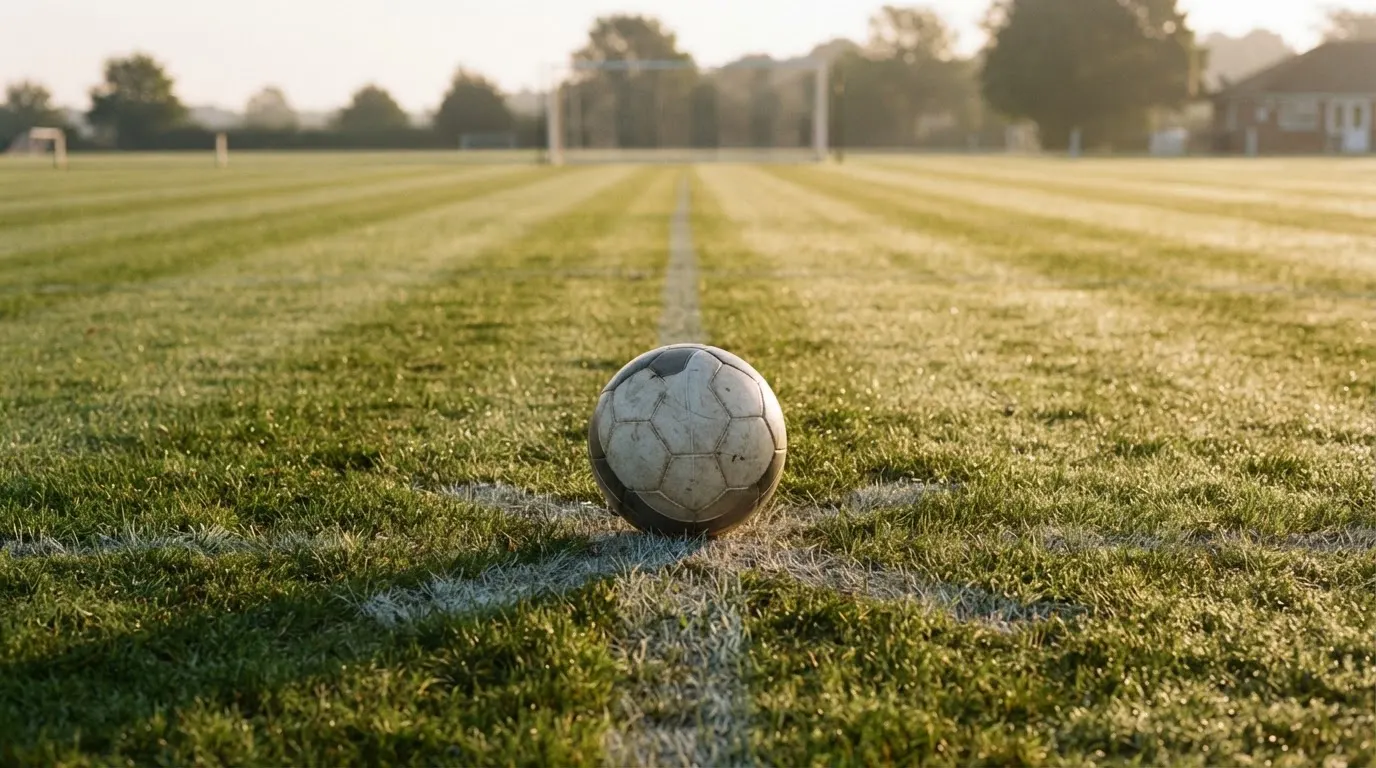 Ein Fußball liegt auf dem Anstoßkreis eines leeren Rasenplatzes im Licht der Morgensonne.