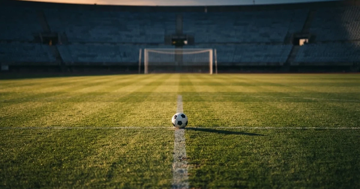 Ein Fußballplatz mit frisch gemähtem Rasen im Abendlicht, ein Ball liegt im Strafraum, im Hintergrund unscharf eine leere Tribüne.