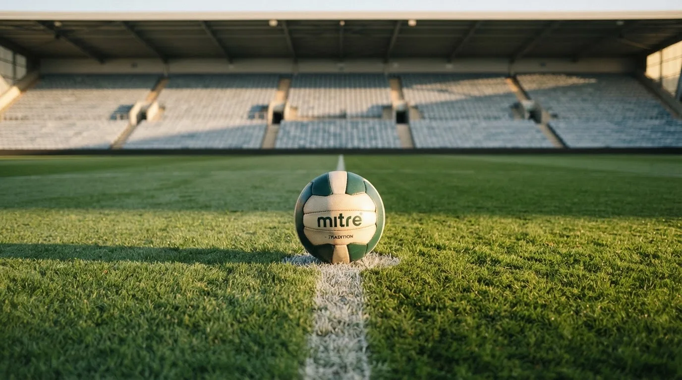 Ein Fußball liegt auf dem Anstoßpunkt eines leeren Rasenplatzes im frühen Morgenlicht, dahinter eine leere Tribüne.