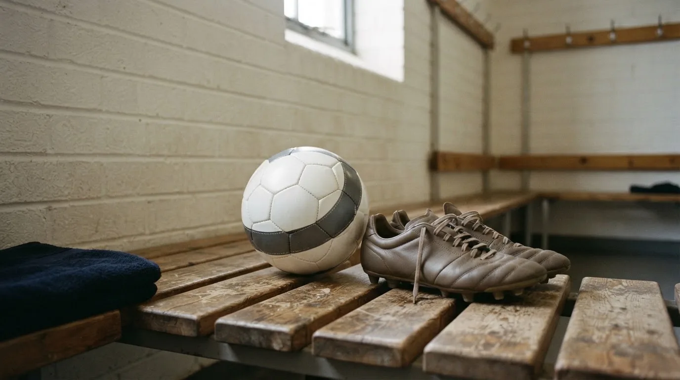 Ein Fußball liegt auf einer Holzbank in einer leeren Umkleide neben einem Paar Fußballschuhen ohne Markenzeichen.