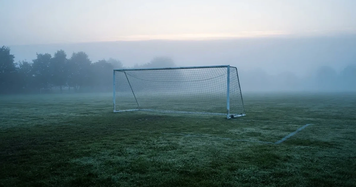 Ein Fußballtor mit Netz steht auf einem leeren Platz im Nebel der Morgendämmerung, der Rasen ist taufeucht.