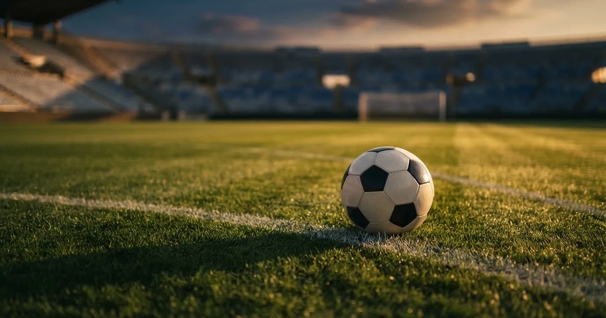 Ein Fußballplatz mit frisch gemähtem Rasen im Abendlicht, ein Ball liegt im Strafraum, im Hintergrund unscharf eine leere Tribüne.