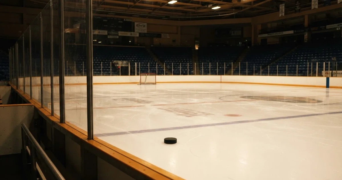 Eine leere Eisfläche in einer Halle mit Banden und Flutlicht, auf dem Eis liegt ein einzelner Puck.