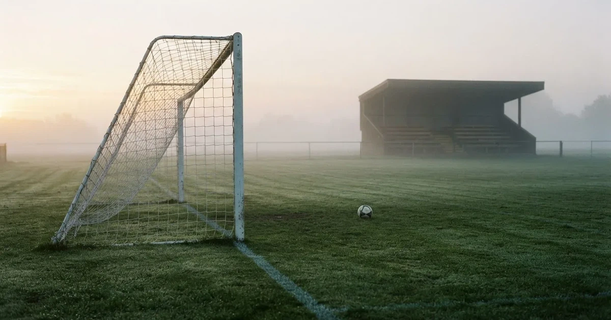 Ein Fußballtor mit Netz steht im Morgennebel auf einem leeren Rasenplatz, im Hintergrund die Silhouette einer kleinen Tribüne.