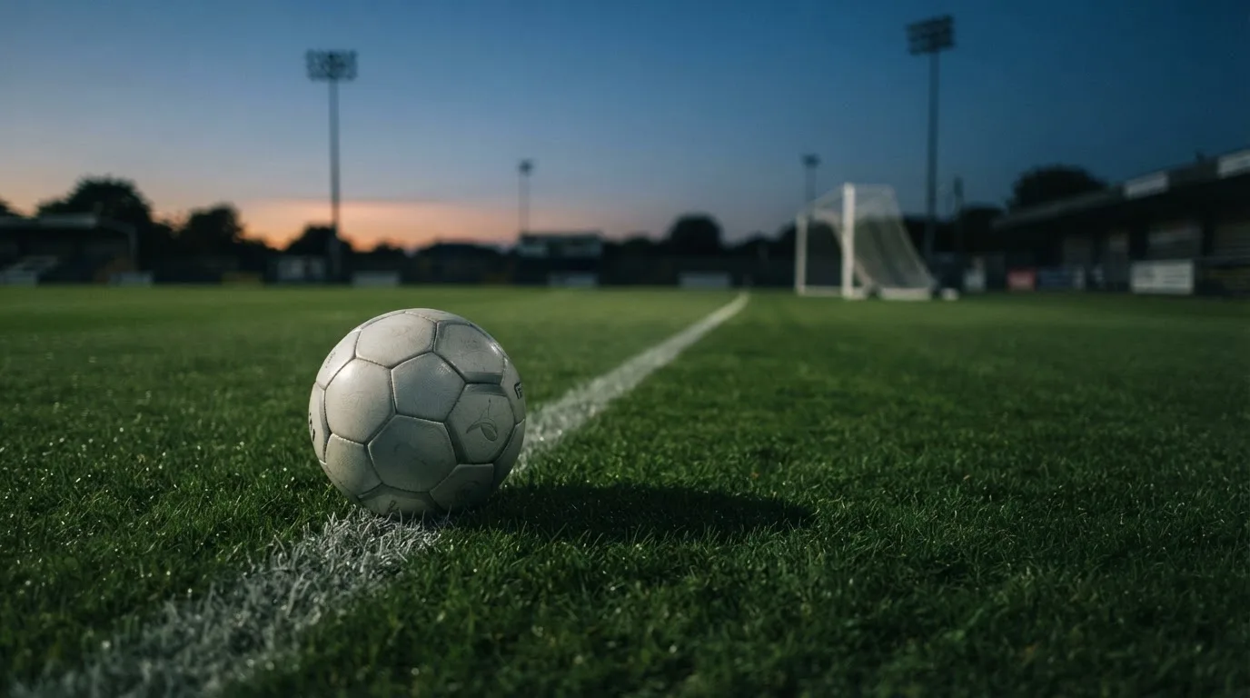 Ein Fußball liegt im Gras eines leeren Platzes, im Hintergrund stehen Flutlichtmasten in der Dämmerung.