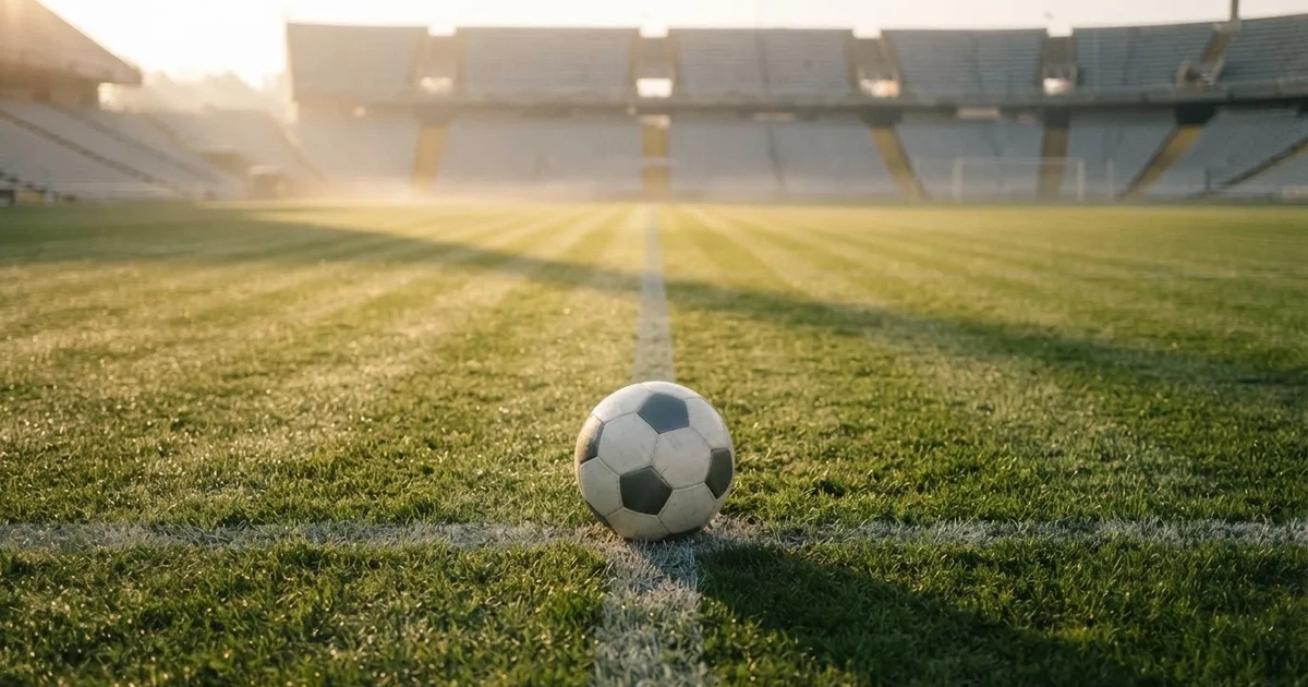 Ein Fußball liegt auf dem Anstoßpunkt eines Rasenplatzes, dahinter leere Tribünen im Licht des frühen Morgens.