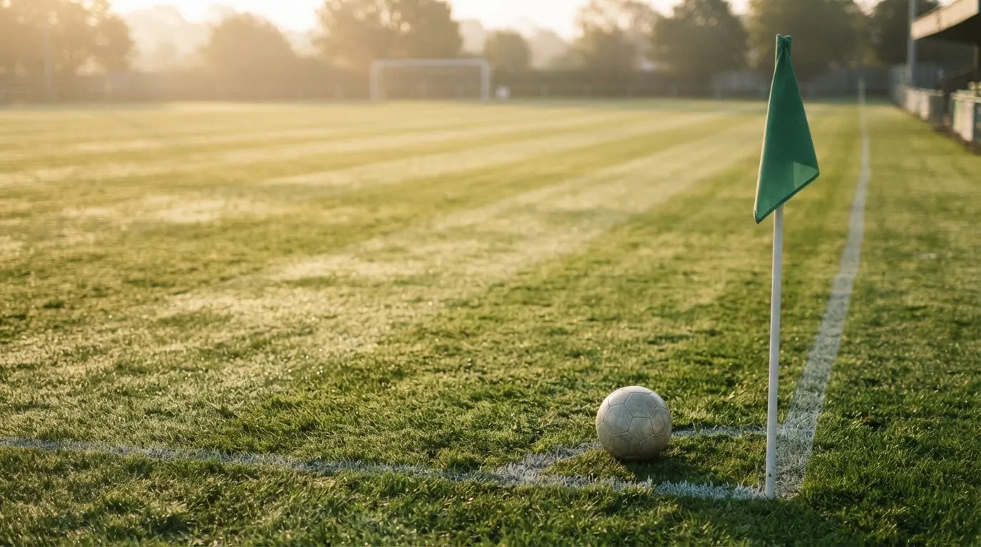 Ein Fußball liegt auf Rasen neben einer Eckfahne ohne Aufdruck auf einem leeren Platz im Morgenlicht.