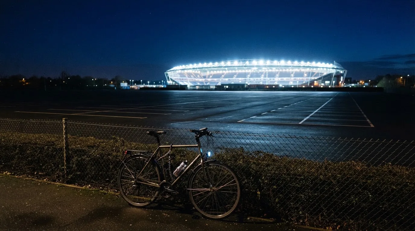 Ein Stadion unter Flutlicht aus der Ferne gesehen, davor ein dunkler Parkplatz und ein einzelnes Fahrrad an einem Zaun.