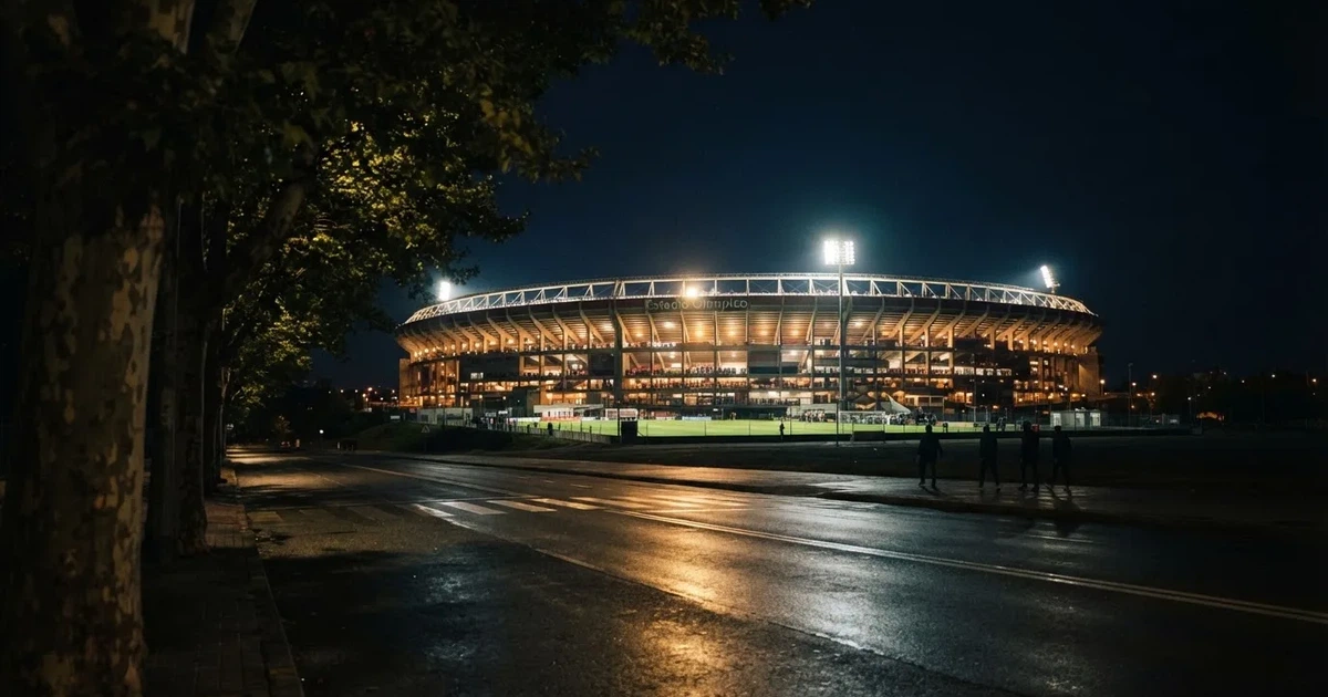 Ein Fußballstadion bei Nacht aus großer Entfernung, hell erleuchtet unter Flutlicht, davor eine dunkle Straße und Bäume.