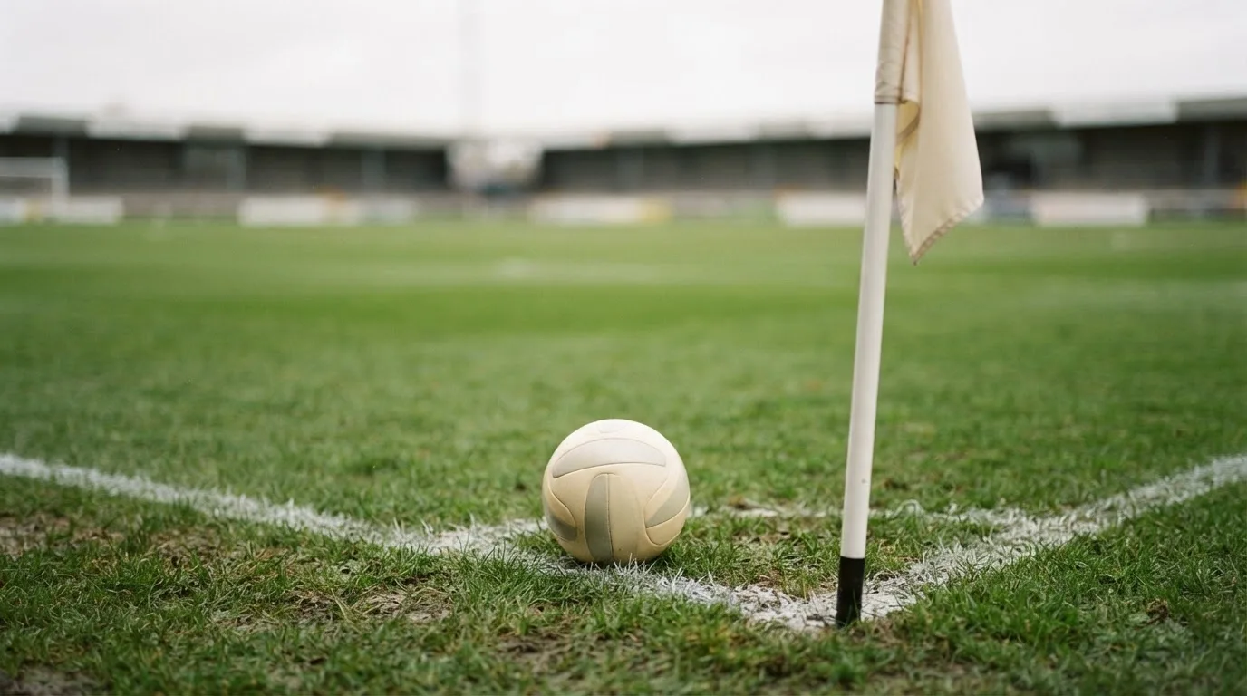 Ein Fußball liegt neben einer Eckfahne auf kurz gemähtem Rasen unter bewölktem Himmel.